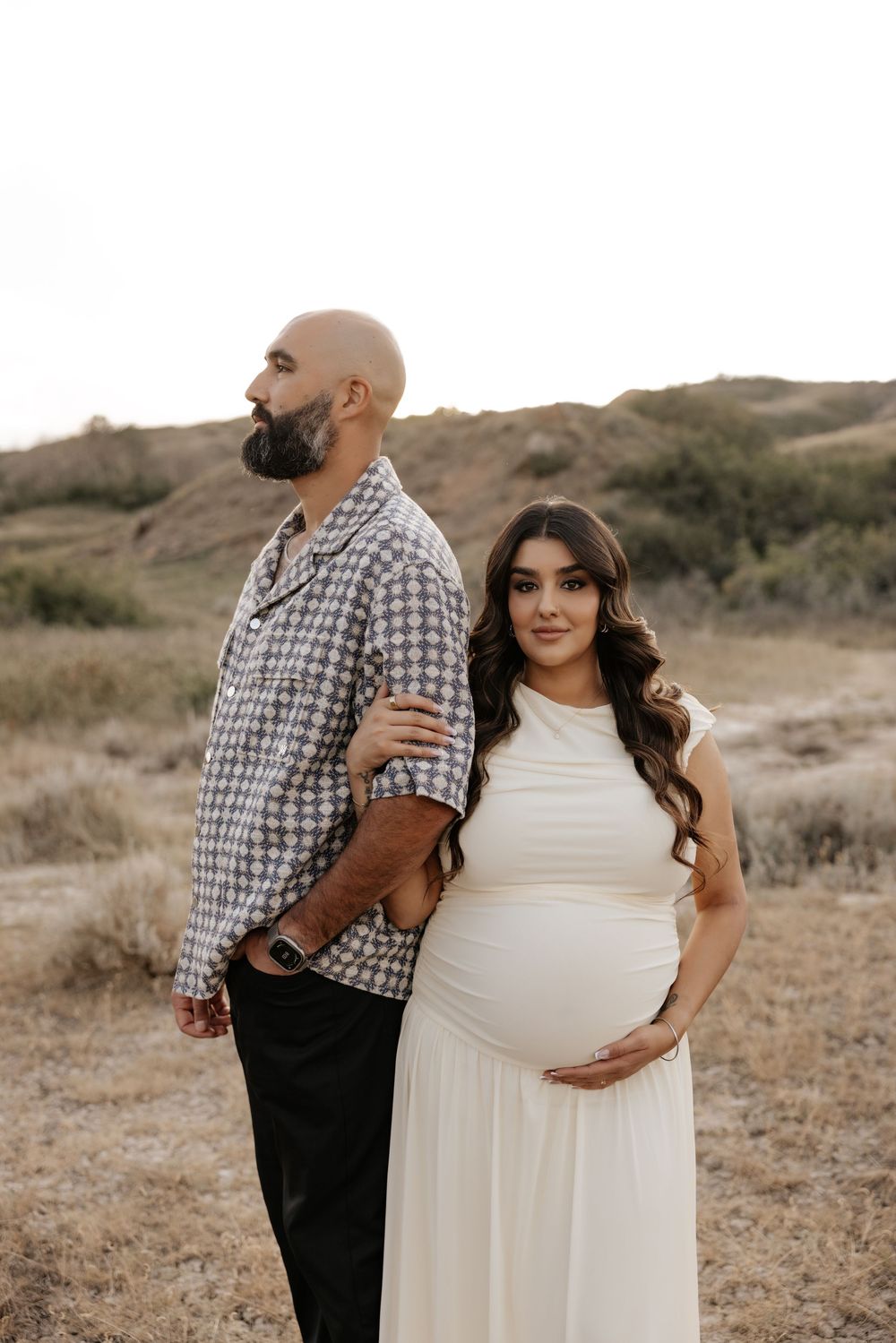 Pregnant woman in white dress with partner standing back-to-back outdoors during golden hour maternity photoshoot.