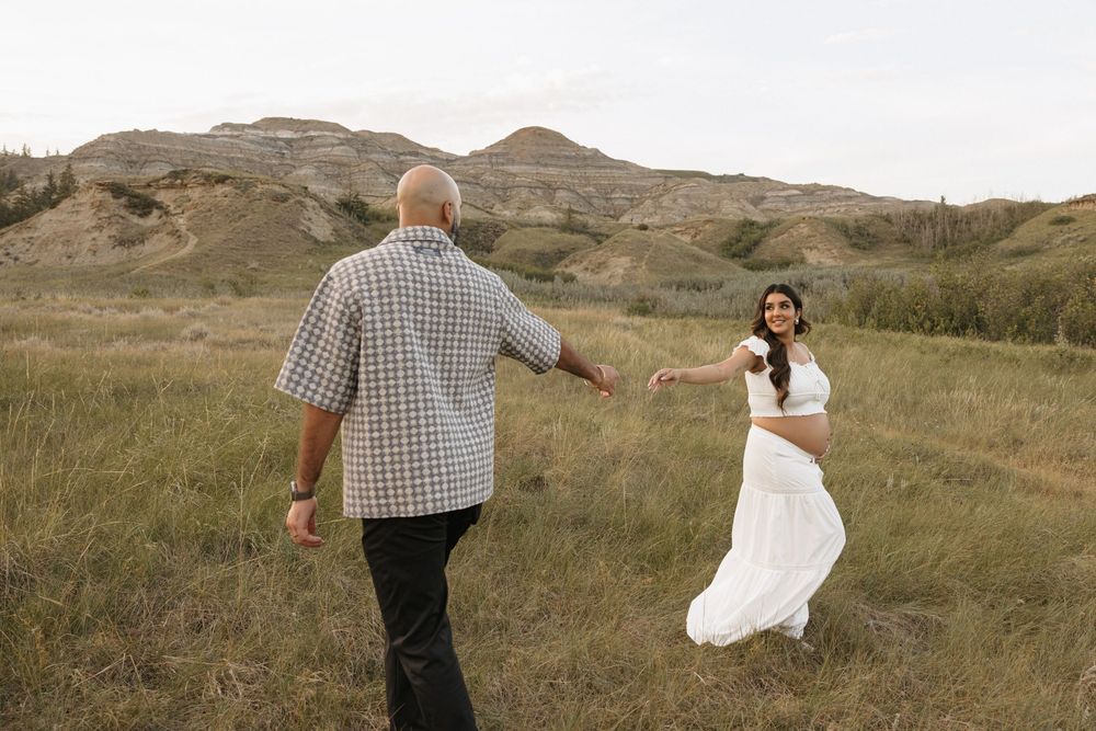 Couple holding hands walking through green meadow with badlands hills in alberta while a photographer captures maternity pictures