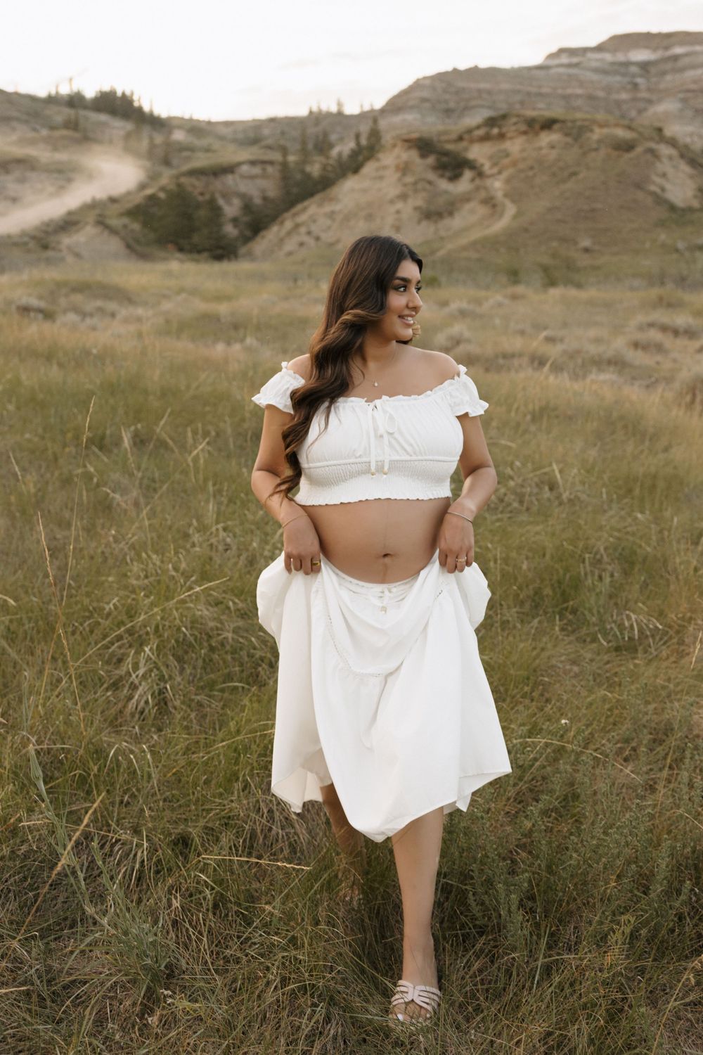Woman in white crop top and skirt posing in a grassy field with badlands hills in the background.