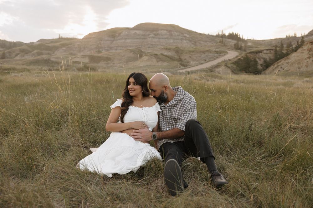 Couple sitting in a grassy field, woman in white dress with baby bump, man embracing her, hills in background.