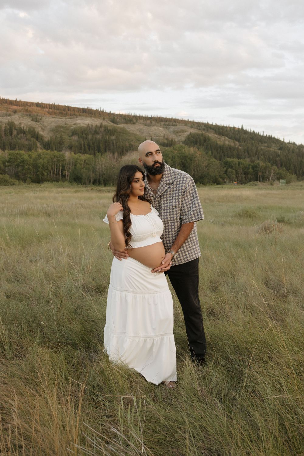 Couple posing in grassy field, man embracing pregnant woman in white dress during maternity photoshoot.