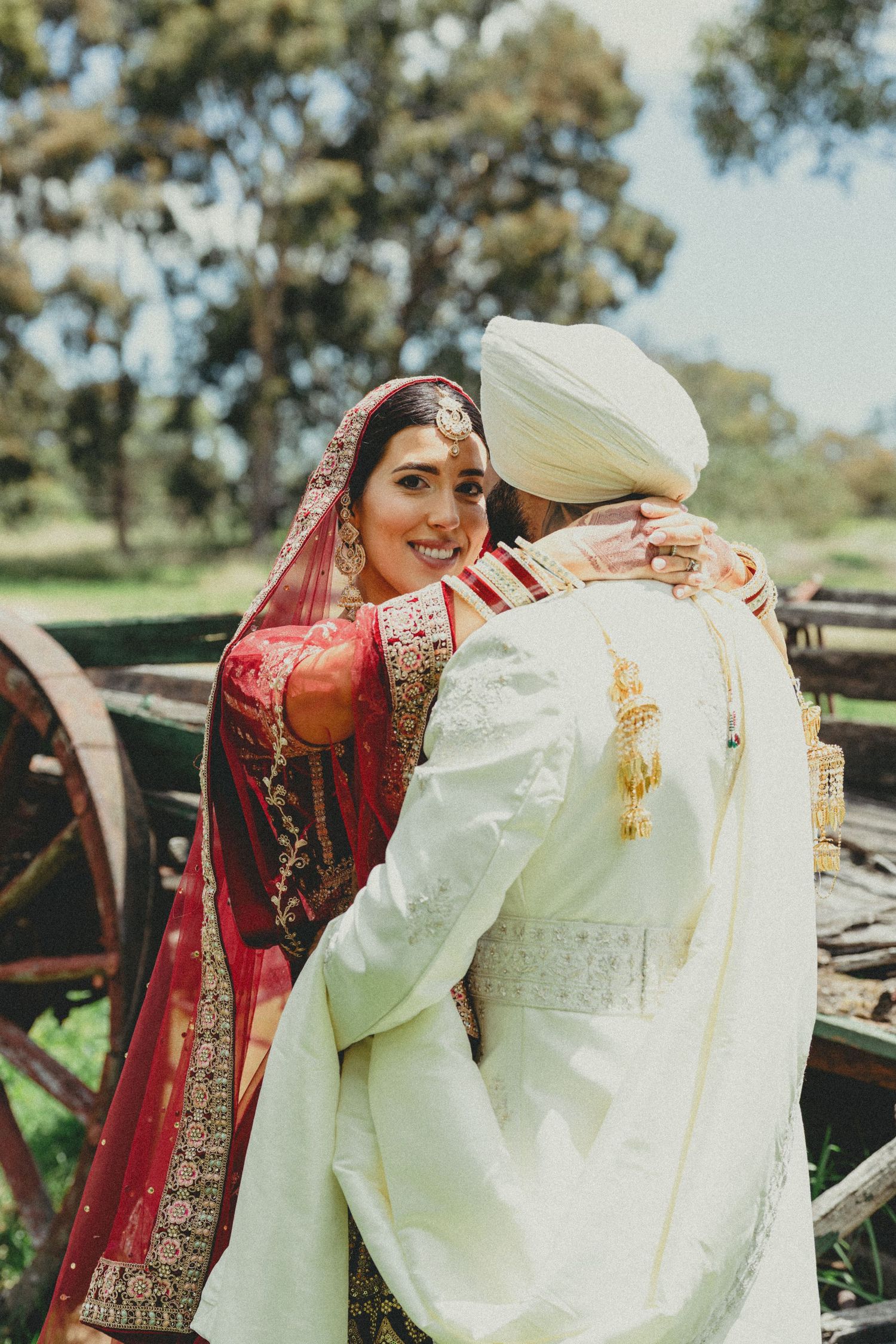 Sikh groom in white turban kissing bride's hand, bride wearing red bridal lehenga and maang tikka at outdoor wedding.