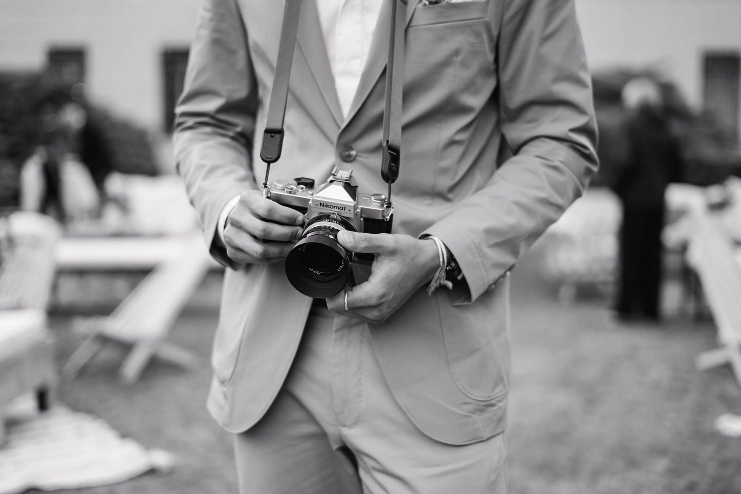 Wedding guest in beige suit holding vintage film camera at outdoor wedding reception, black and white photo.