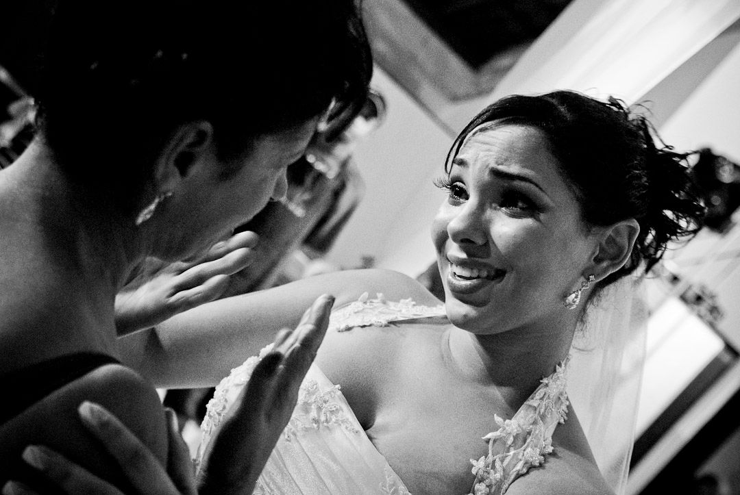 Black and white wedding photo of a smiling bride in a tiara holding bouquet, surrounded by guests outdoors.