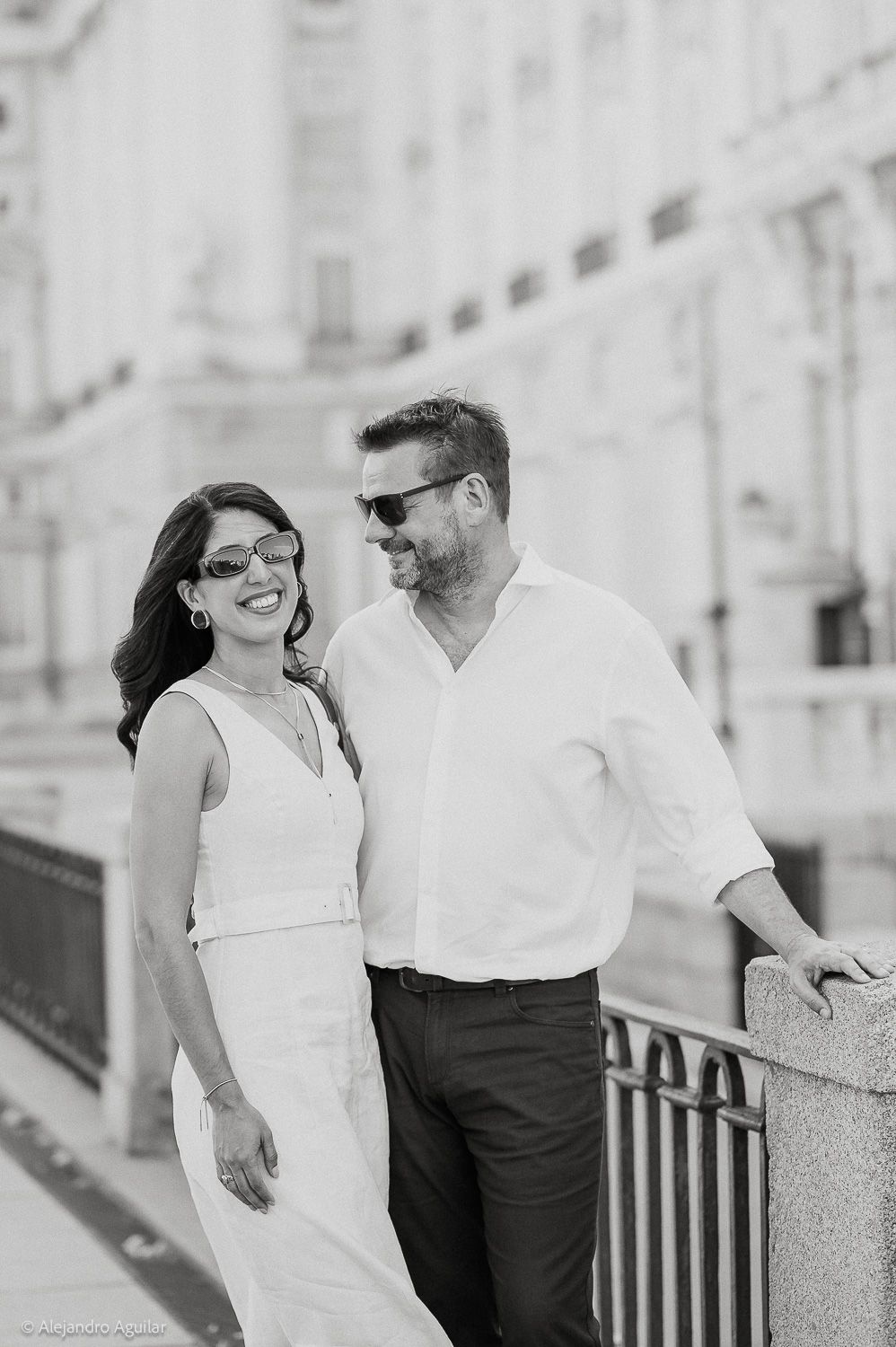Black and white photo of a smiling couple in sunglasses posing outdoors near a railing with a grand building behind them.