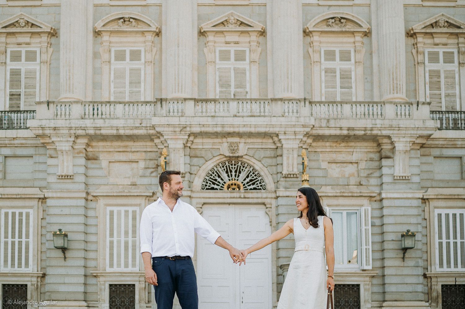 Couple holding hands in front of an elegant white neoclassical palace building during a romantic outdoor photoshoot.