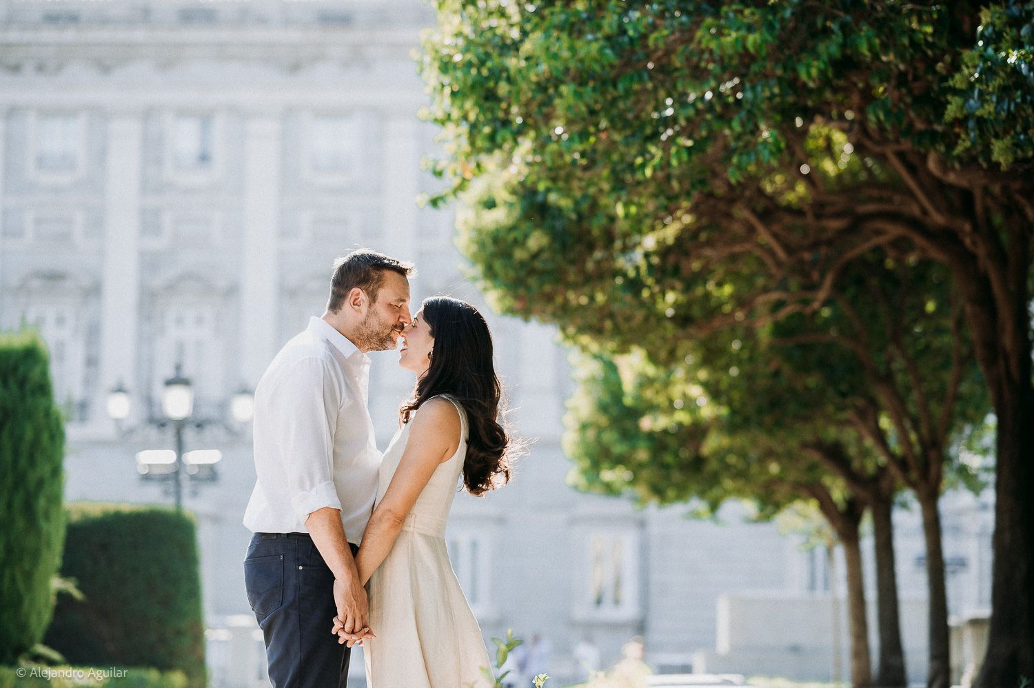 Couple sharing a romantic kiss outdoors near ornate building and lush green trees on a sunny day.