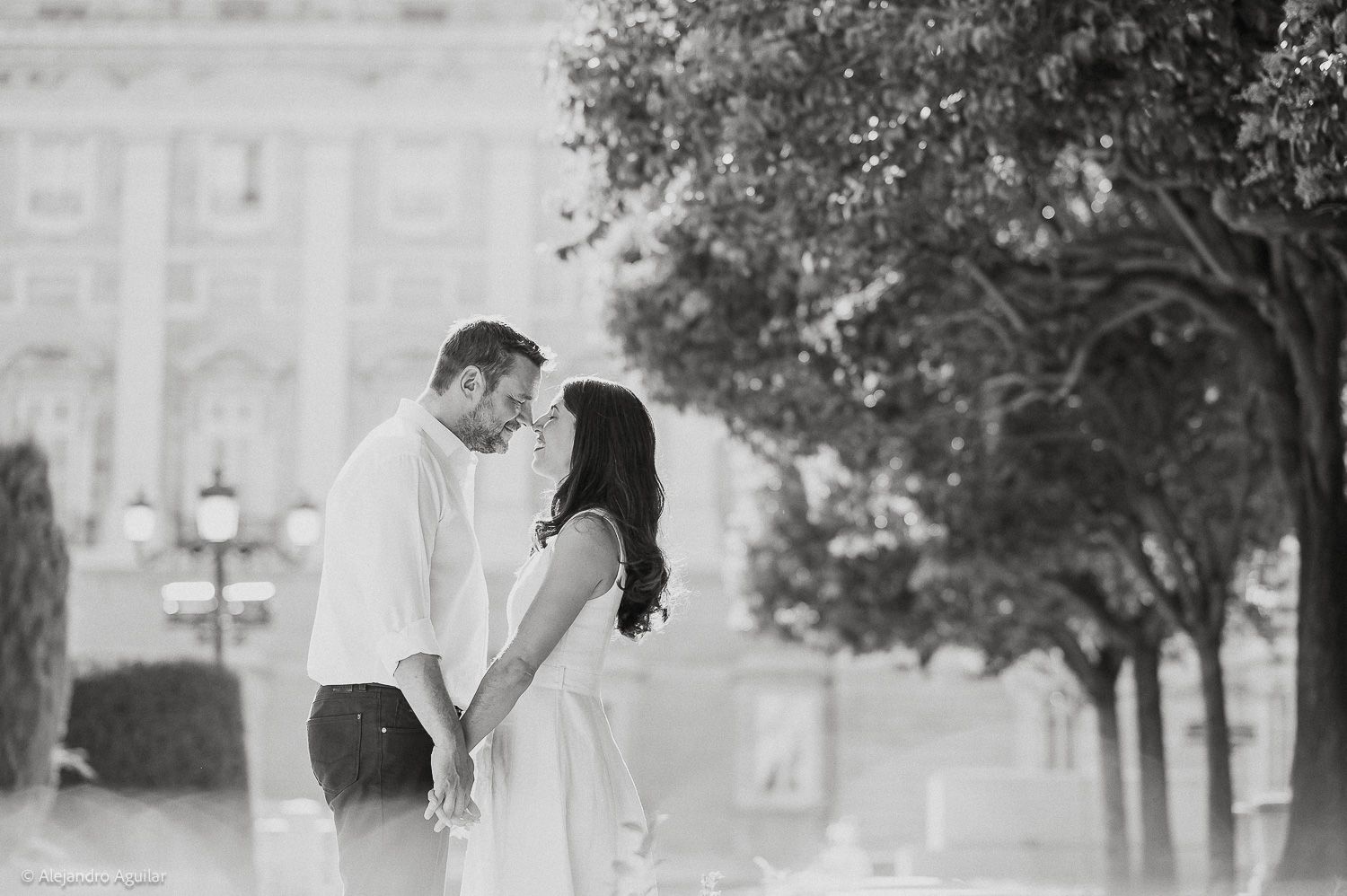 Black and white photo of a romantic couple holding hands and leaning in for a kiss outdoors near trees.