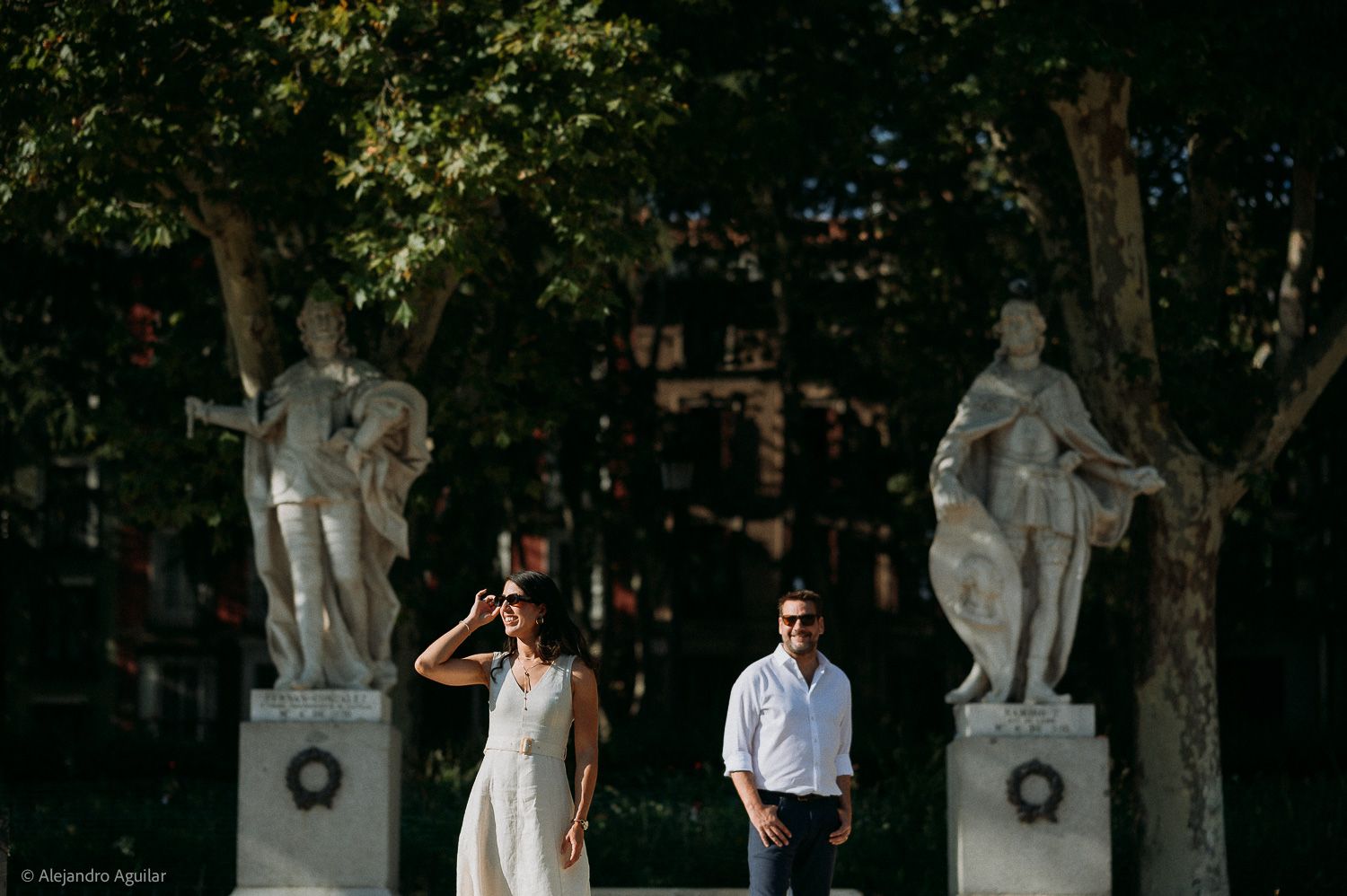 Couple posing between classical statues in a sunlit park, woman in white dress, man in blue shirt.