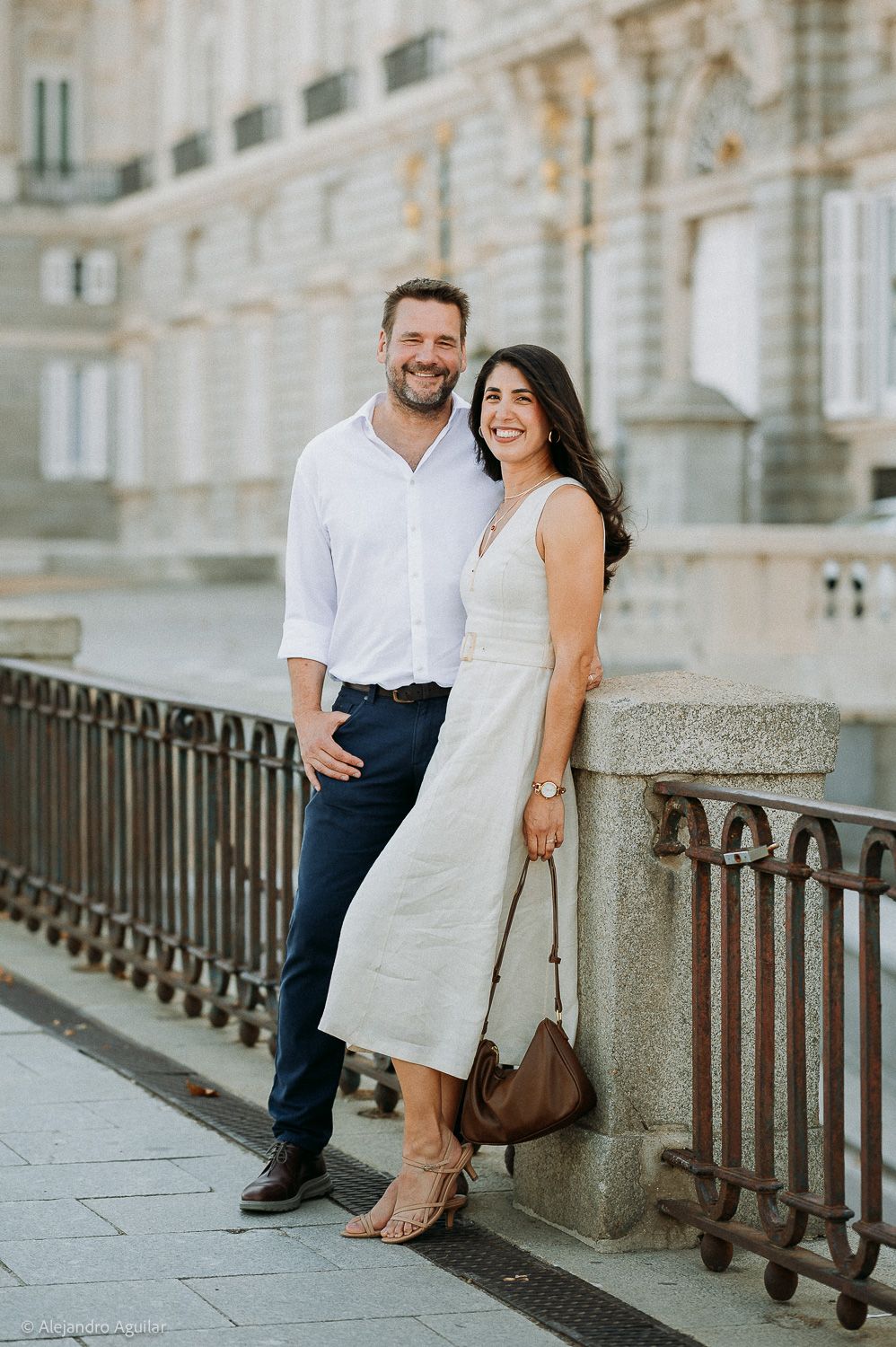 Happy couple posing together on a cobblestone walkway near ornate iron railings and a grand historic building.
