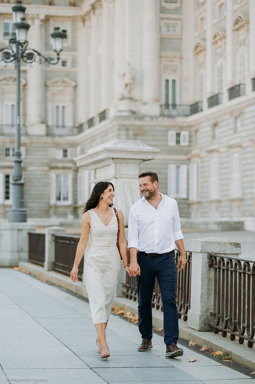 A couple walks hand-in-hand smiling near a grand European palace, woman in white jumpsuit, man in white shirt.