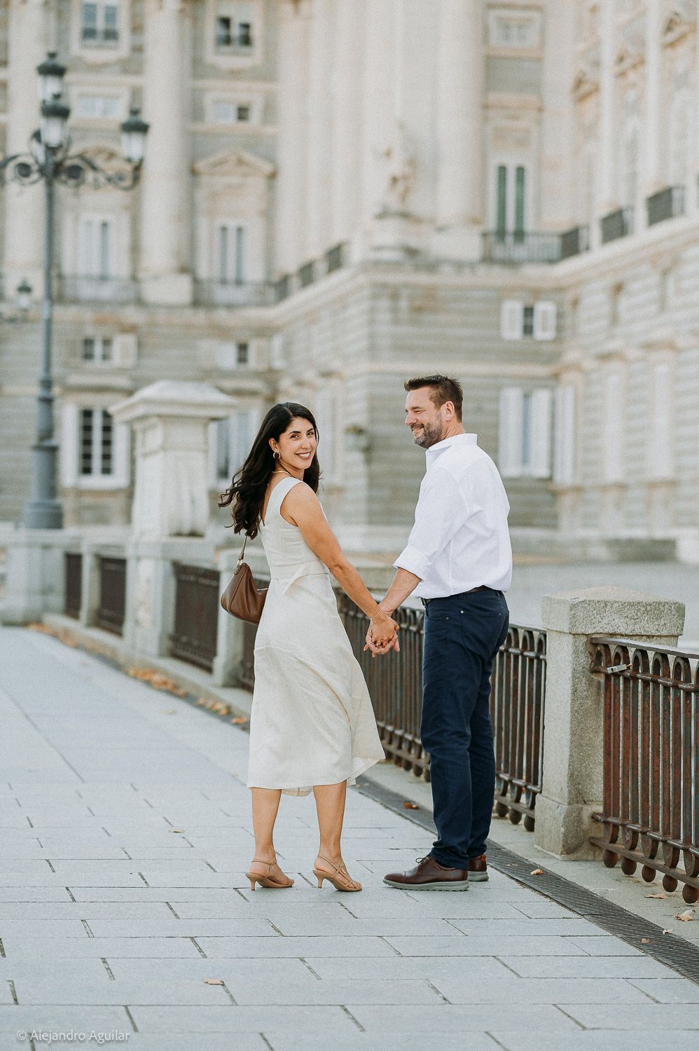 Couple holding hands and smiling in front of a grand European building, woman in beige dress, man in white shirt.