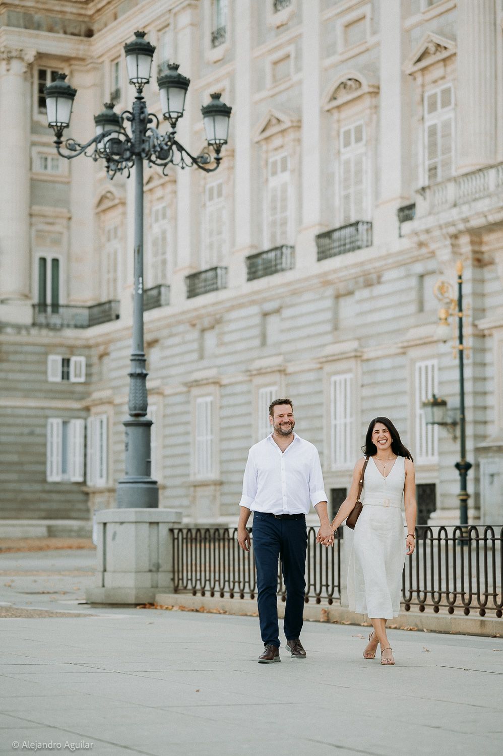 Couple walking hand in hand near the Royal Palace of Madrid, dressed in white and navy outfits.