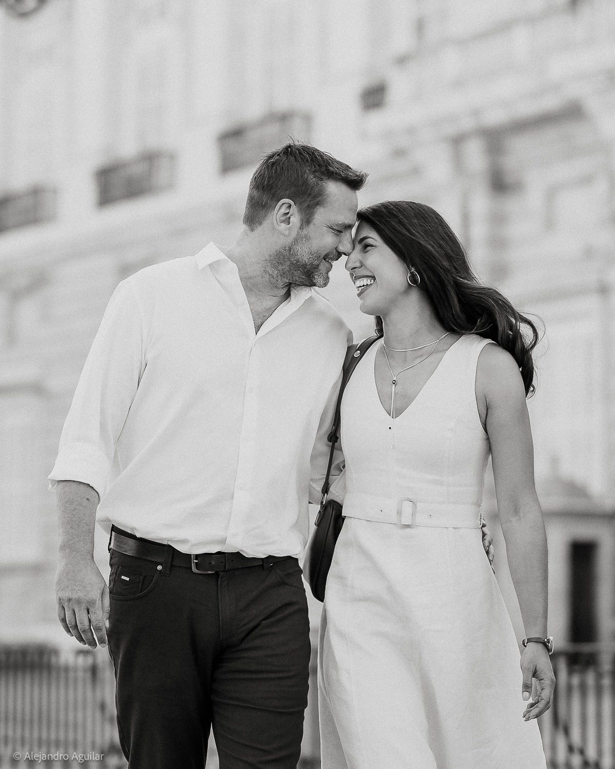 Black and white photo of a smiling couple in white outfits sharing a romantic moment outdoors near a classic building.