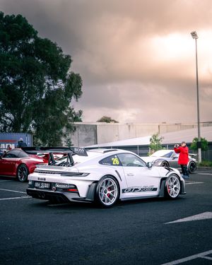 White Porsche 911 GT3 race car number 26 with BOSS sponsorship in parking lot under dramatic cloudy sky.