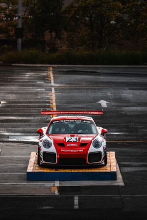 Red and white Porsche 911 GT2 RS race car number 23 on display platform in wet parking lot.