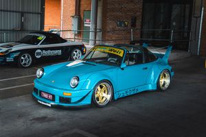 Teal RWB wide-body Porsche 993 with gold wheels and yellow livery parked outside brick building.