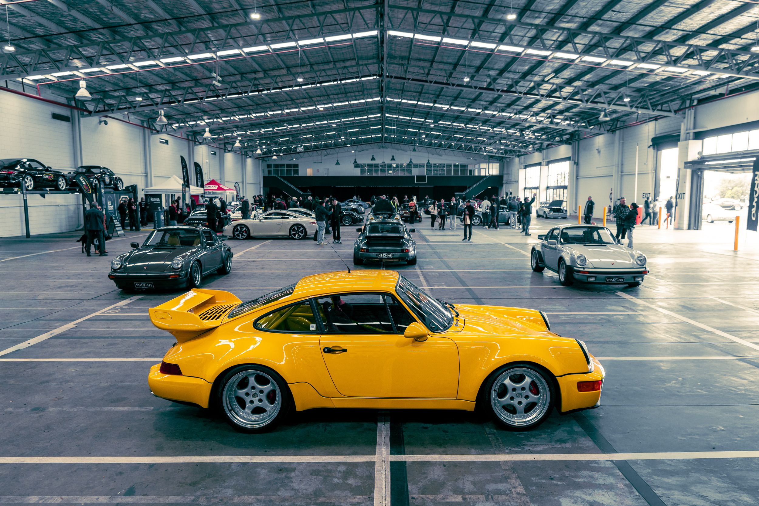 Yellow Porsche 911 964 displayed in a large warehouse surrounded by other classic Porsche models.