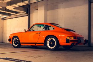 Rear three-quarter view of a classic orange Porsche 911 parked in a sunlit concrete garage.