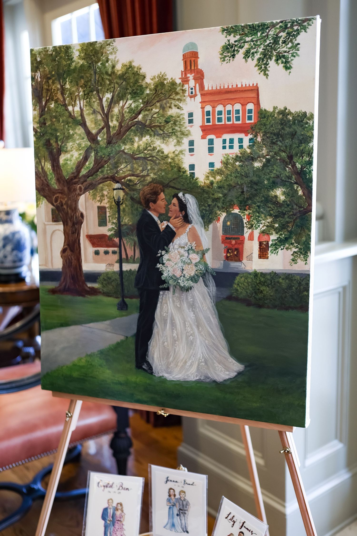 A wedding painting on an easel depicting a bride and groom embracing in front of a red brick building surrounded by green trees.