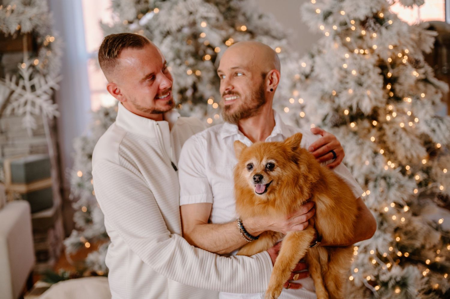 Two men in white outfits holding a fluffy Pomeranian dog in front of decorated Christmas trees with warm lights.