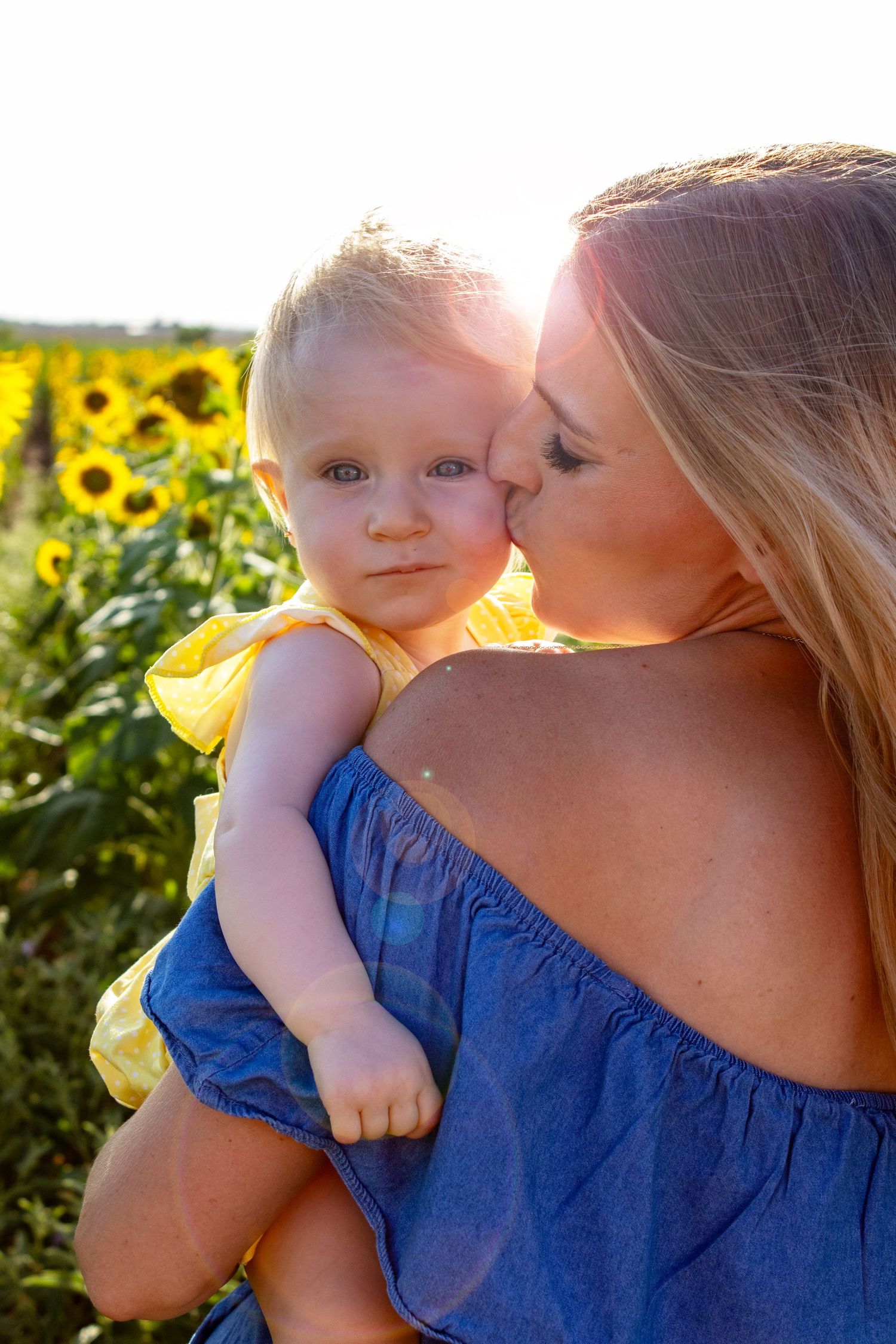 A mother in a blue dress kisses her baby girl in a yellow dress in a sunflower field.