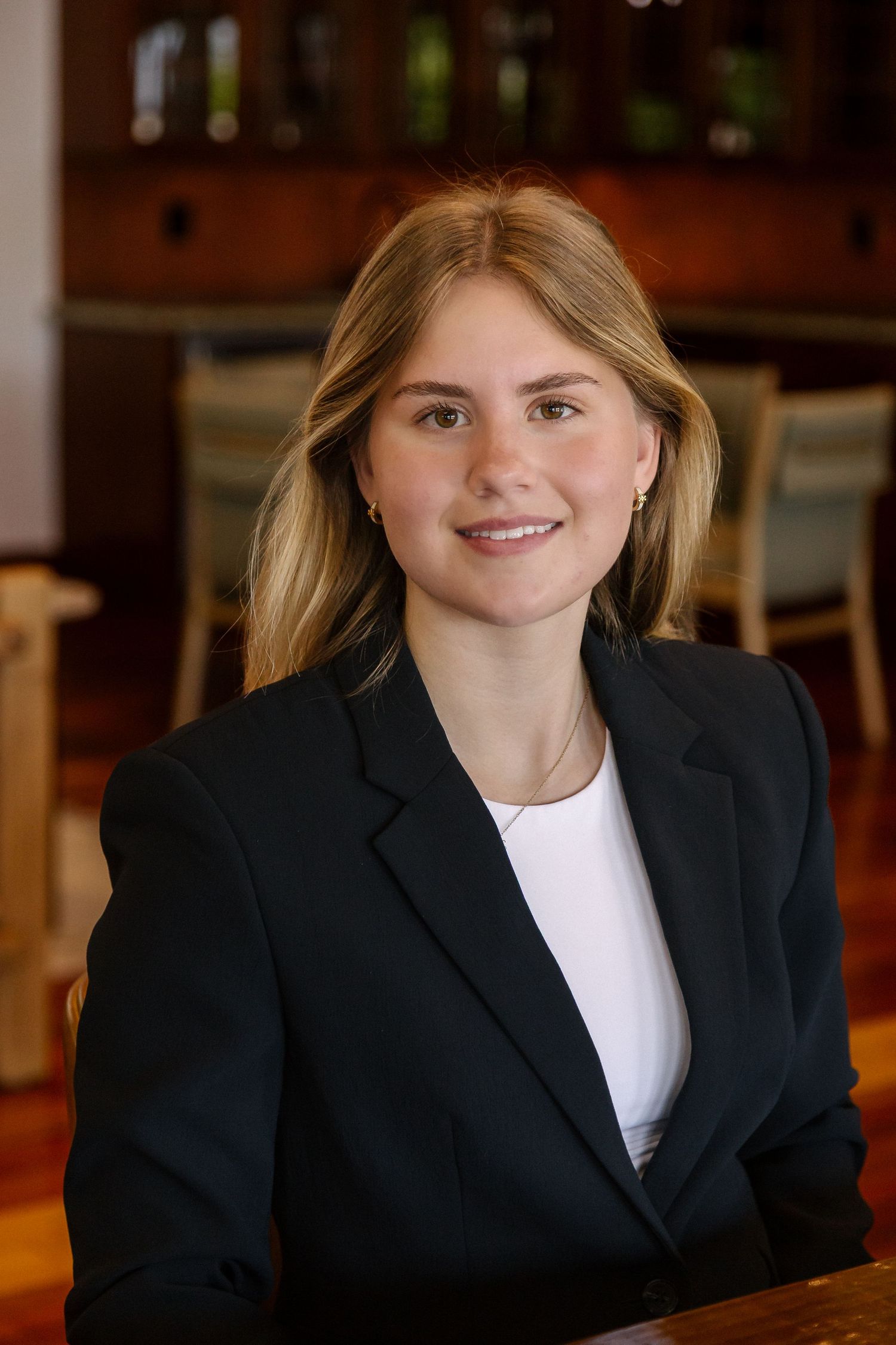 Young professional woman in black blazer and white top smiling confidently in a modern restaurant setting.