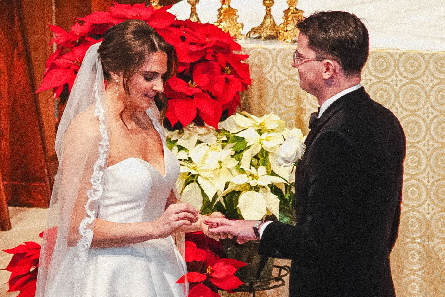 Bride and groom exchange rings at altar surrounded by red and white poinsettias at Christmas wedding.
