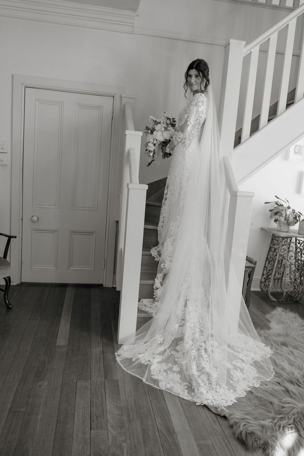 Bride in lace wedding gown with cathedral veil holds bouquet on staircase in black and white portrait.  Clifton Woolshed