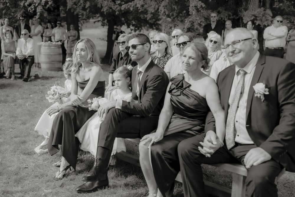 Black and white photo of wedding guests seated on benches outdoors, smiling during a ceremony.  Clifton Woolshed