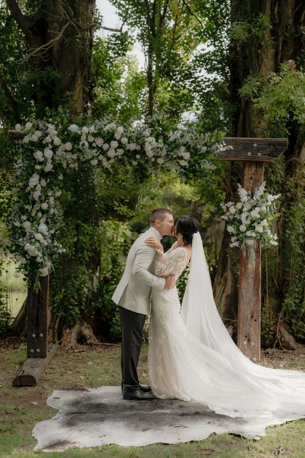 Bride and groom share first kiss under rustic wooden arch adorned with white flowers surrounded by lush green trees.  Clifton Woolshed