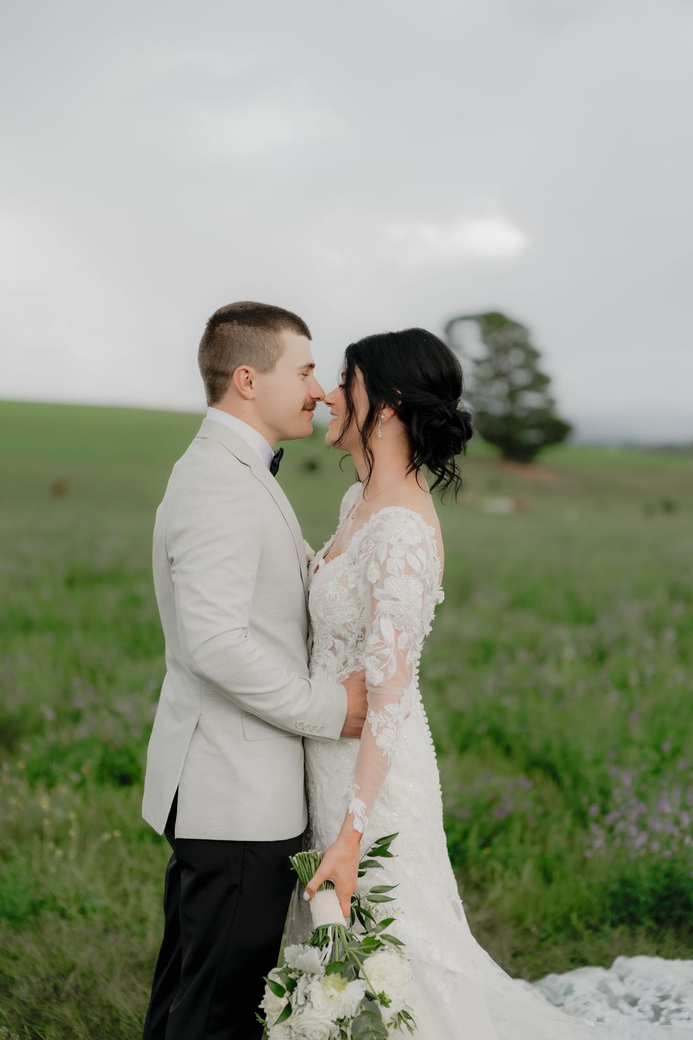 A bride and groom share a romantic moment in a lush green field, with the bride holding a white bouquet.  Clifton Woolshed