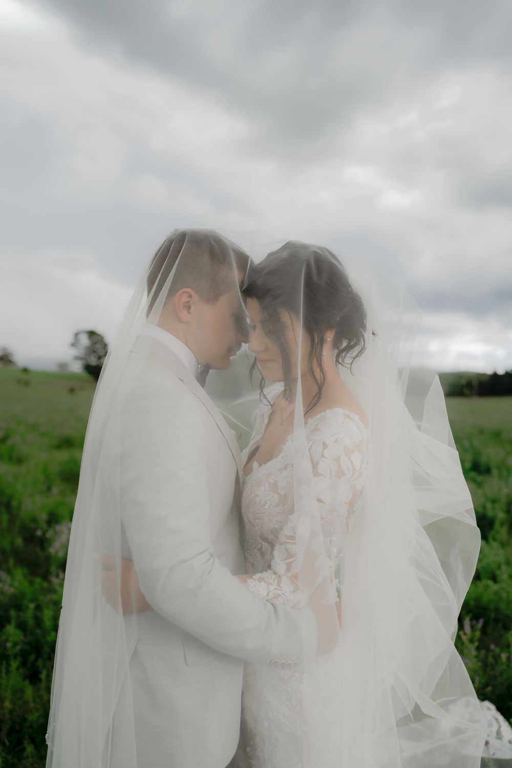 Bride and groom share an intimate moment under a flowing veil in a green field with overcast skies. Clifton Woolshed