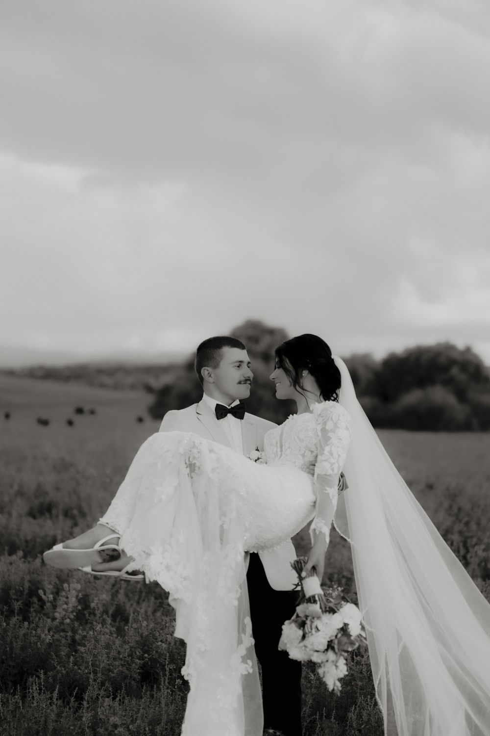 Black and white photo of groom carrying bride in lace wedding dress and veil in a scenic countryside field.  Clifton Woolshed