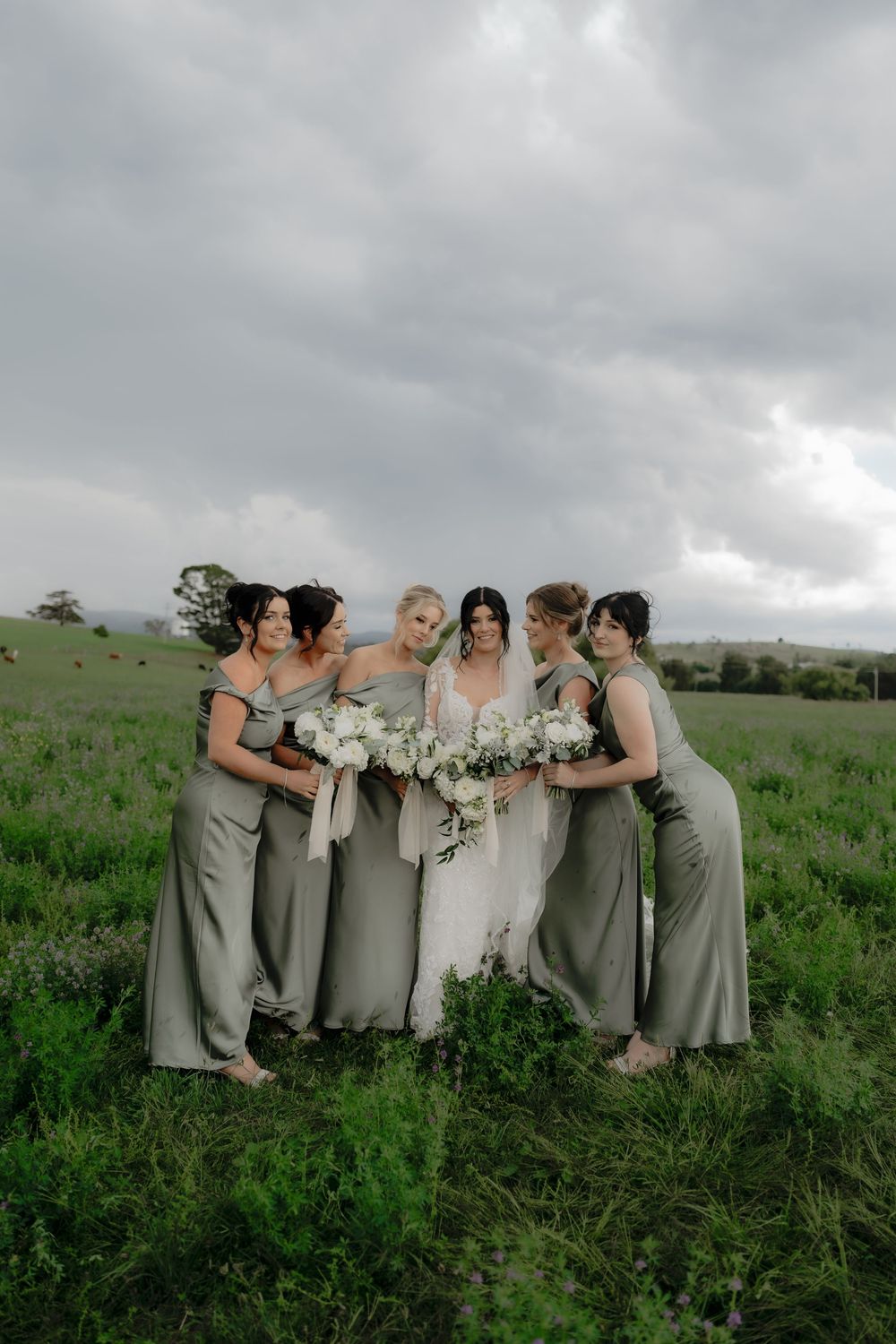 Bride in white gown with five bridesmaids in sage green dresses holding white bouquets in a lush green field.  Clifton Woolshed