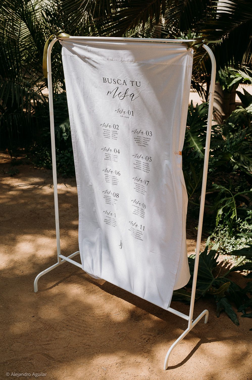 Elegant white fabric seating chart sign reading Busca Tu Mesa displayed on a gold rack at an outdoor wedding.