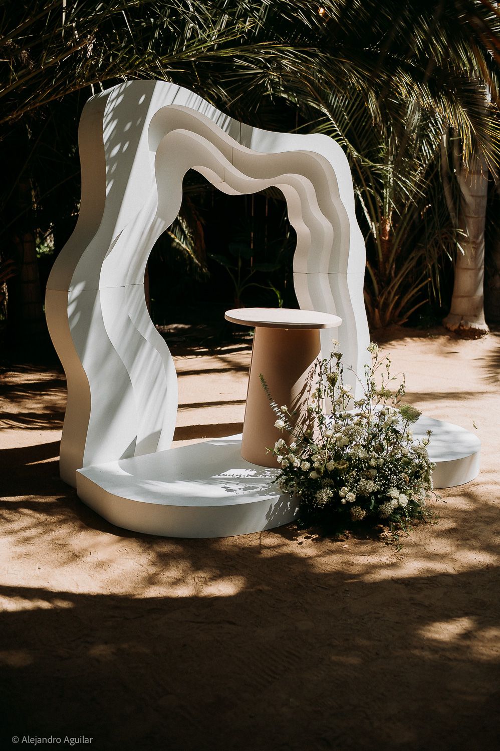 White wavy arch wedding ceremony backdrop with floral arrangement and pedestal table, set outdoors under palm trees.