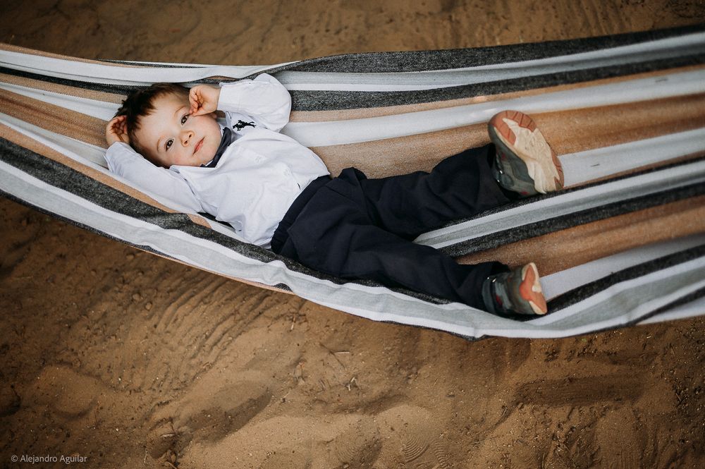 Young boy in formal wear relaxing in a striped hammock over sandy ground, smiling contentedly.
