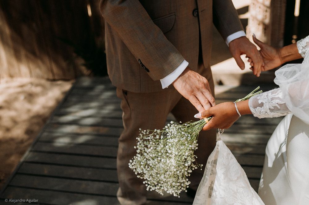 Bride holding baby's breath bouquet with groom in brown suit on wooden bridge at outdoor wedding.