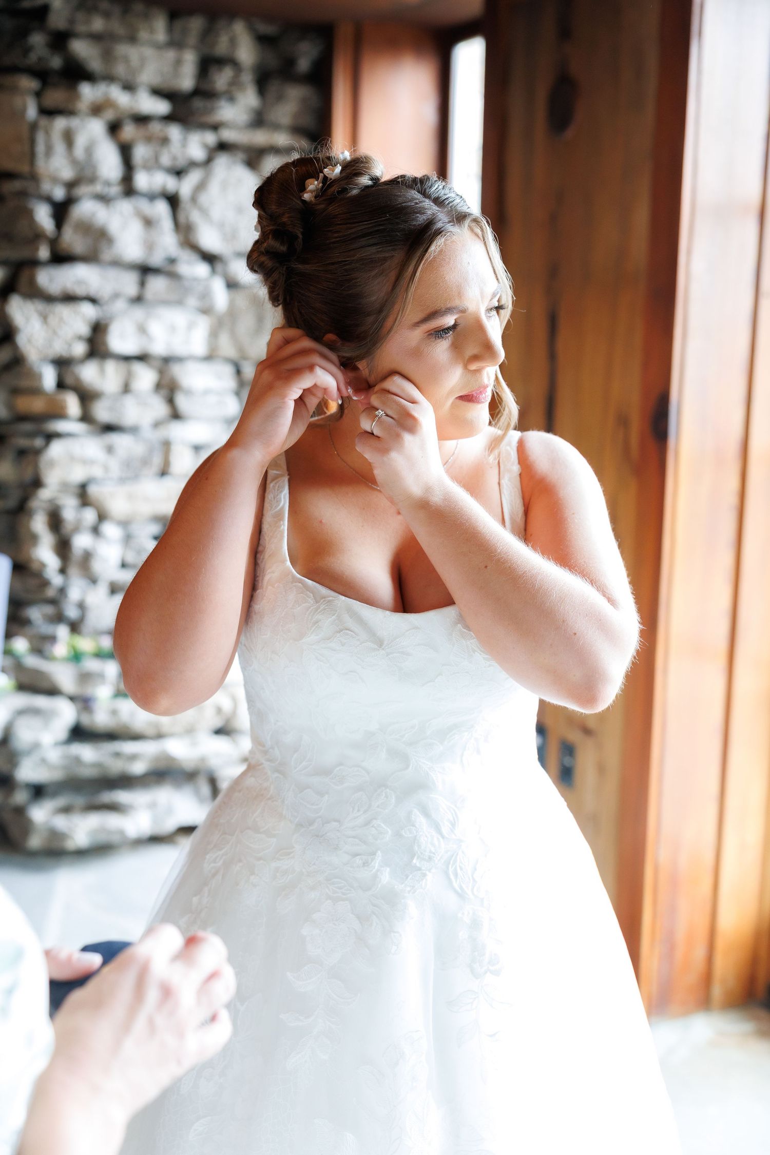 Bride in white lace wedding gown putting on earrings near rustic stone wall and wooden door.