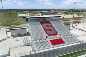Aerial view of a high school football stadium with red S logo on white bleachers, surrounded by green fields.