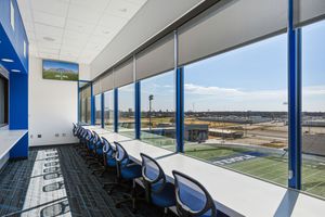 Modern press box interior with long desk, blue chairs, and panoramic windows overlooking the football field.