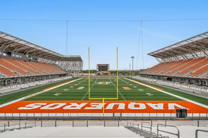 Vaqueros football stadium with bright orange turf, yellow goalposts, and tiered seating under a clear blue sky.