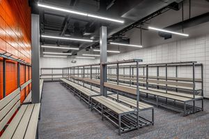 Spacious team locker room with rows of metal benches, shelving units, orange accents, and bright overhead lighting.