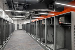 Long hallway of individual metal mesh lockers with orange accents in a modern sports facility locker room.