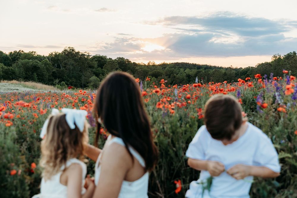 Three people in white dresses stand in a vibrant poppy field at sunset with colorful wildflowers and trees.