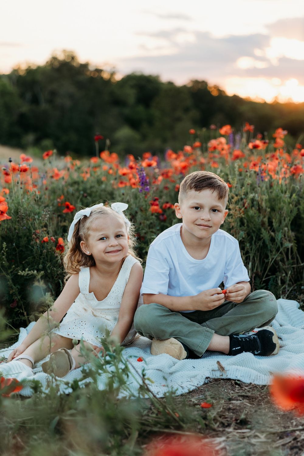 Two children sitting on a blanket in a vibrant poppy field at sunset, smiling at the camera.