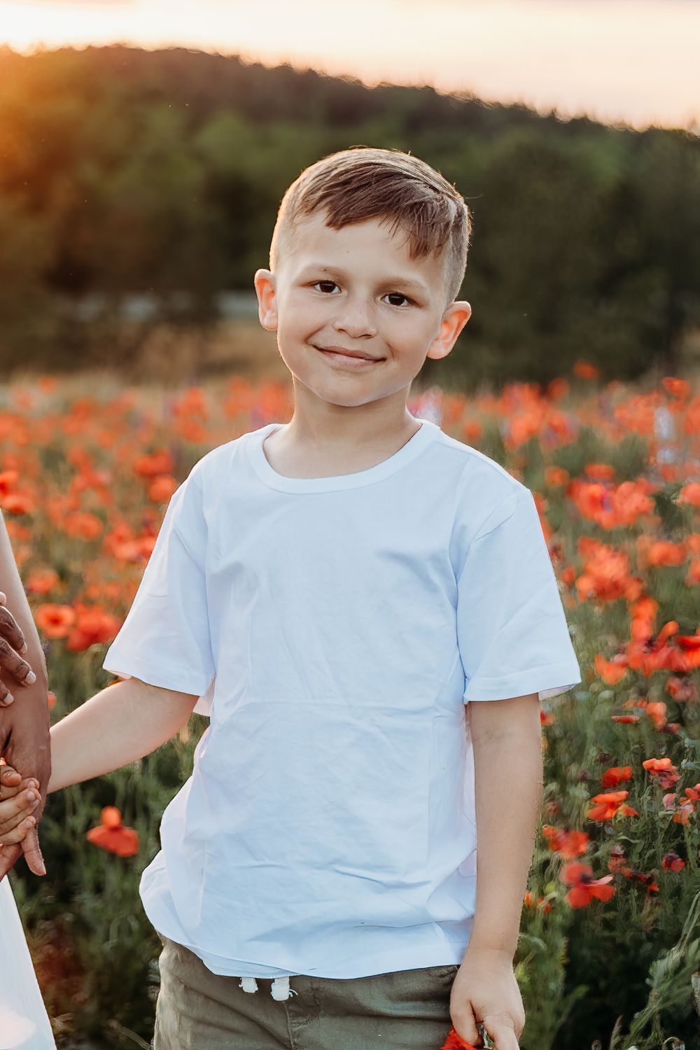 Smiling boy in white t-shirt standing in a red poppy field at golden hour sunset.