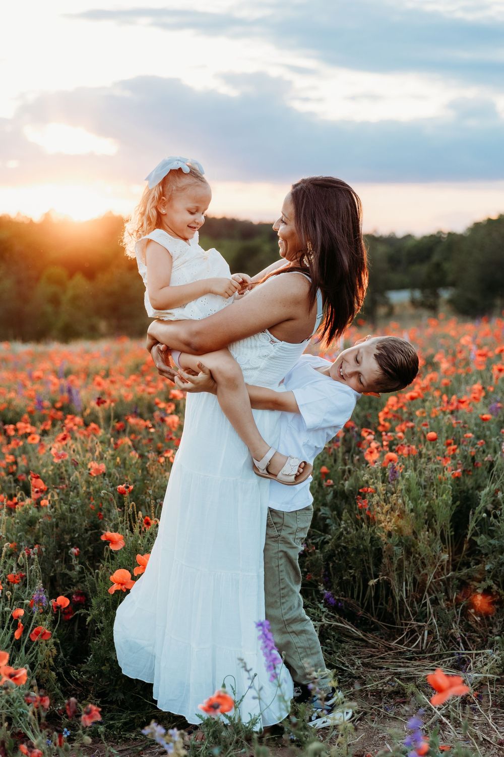 Mother in white dress holds daughter while son hugs her in a vibrant poppy field at golden sunset.