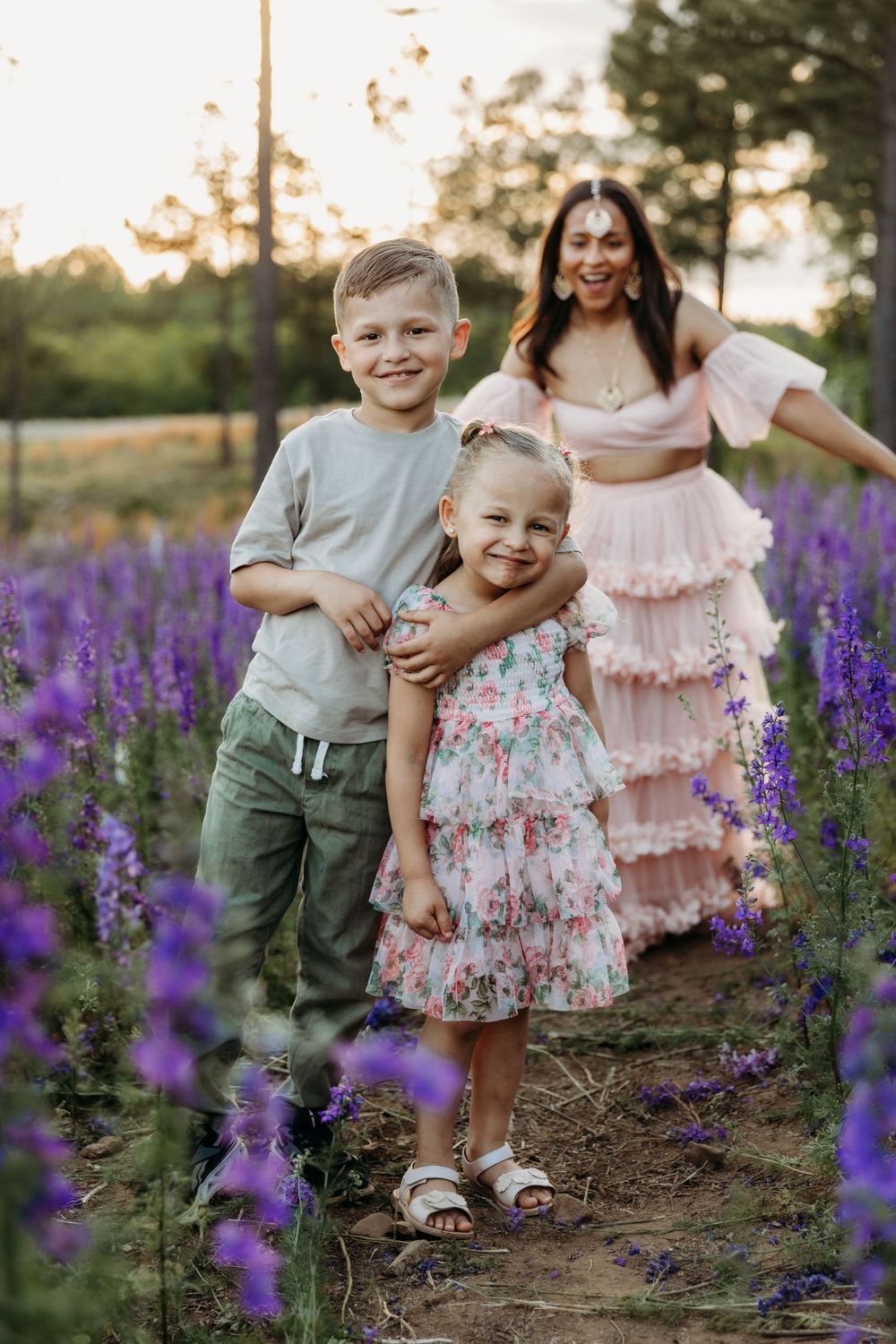 A mother in a pink dress poses with her two young children in a purple wildflower field at golden hour.